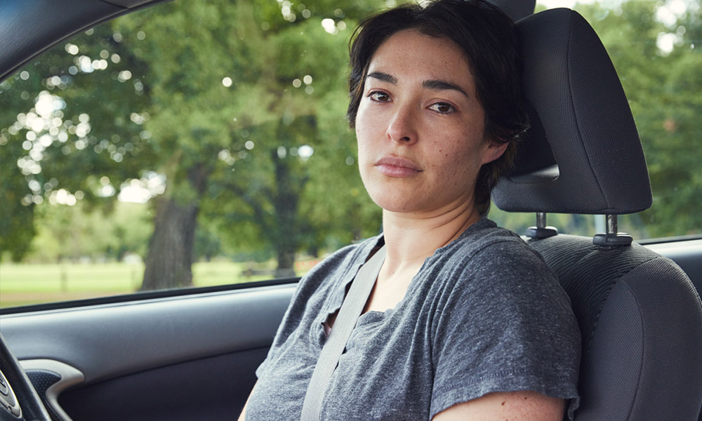 Young mother sits in a car with tears in her eyes looking towards the camera.